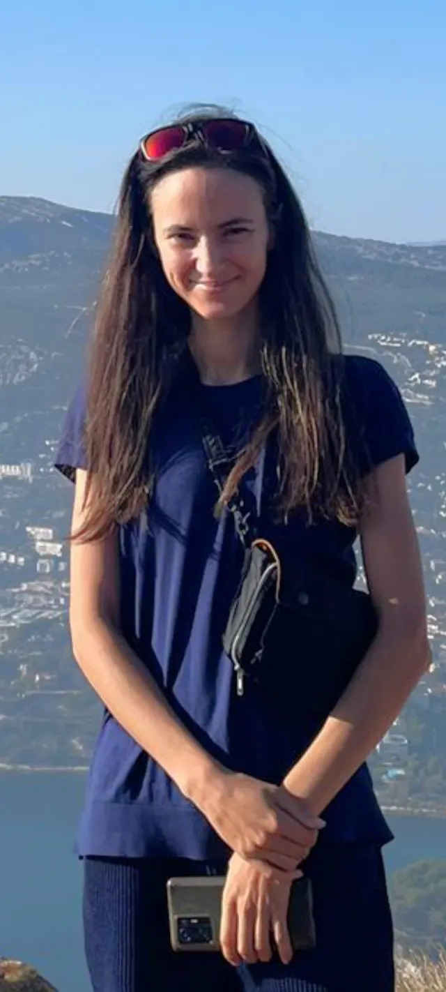 Sabrina Smirnova, debout en extérieur devant une baie bordée de collines : longs cheveux bruns, lunettes de soleil relevées sur la tête, t-shirt bleu marine, les mains posées devant elle.